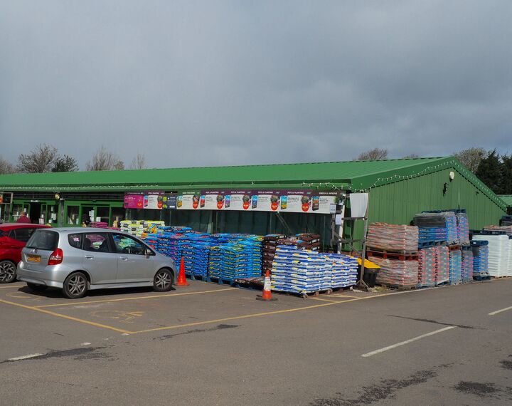 exterior view of a large green shed-like building, with bags of compost in front, and two parked cars