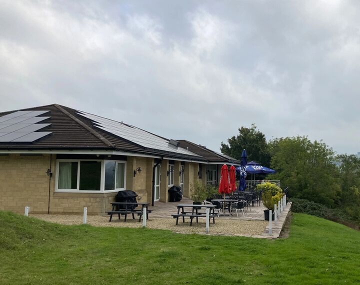 Golf course club house exterior with solar panels on roof and with umbrellas on patio and a view of hills behind 