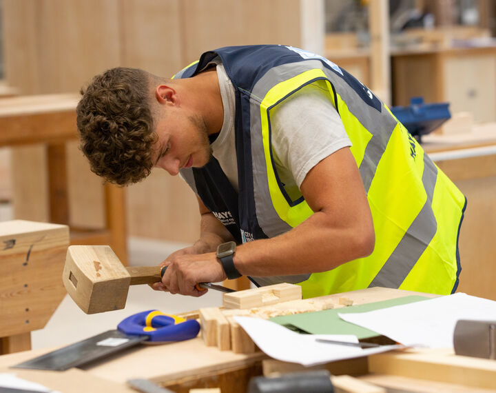 A young man is in a workroom doing woodwork with a wooden hammer, wearing a high-vis yellow jacket