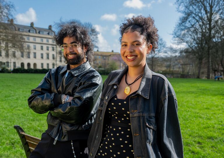 two young people leaning against a park bench, smiling at the camera