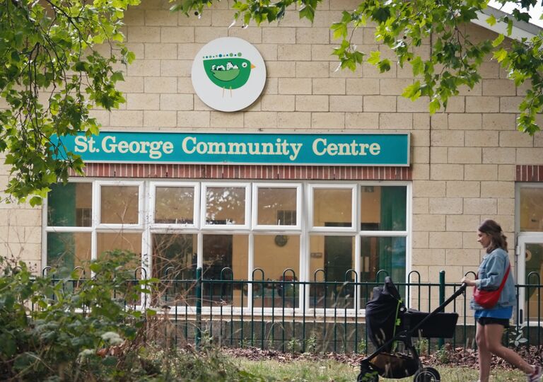 a young mother pushing a pushchair in front of a yellow brick building, with a teal sign that says 'St George Community Centre' in white writing.