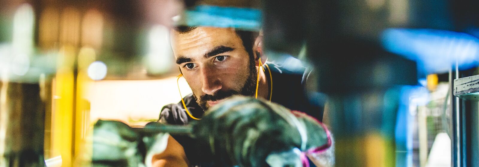 A man is holding a tool in his hands, while wearing gloves. He is looking intently and wearing earplugs connected on a wire. The area around him is blurry, so we cannot see what he is working on, but we can tell it is a manufacturing site. 