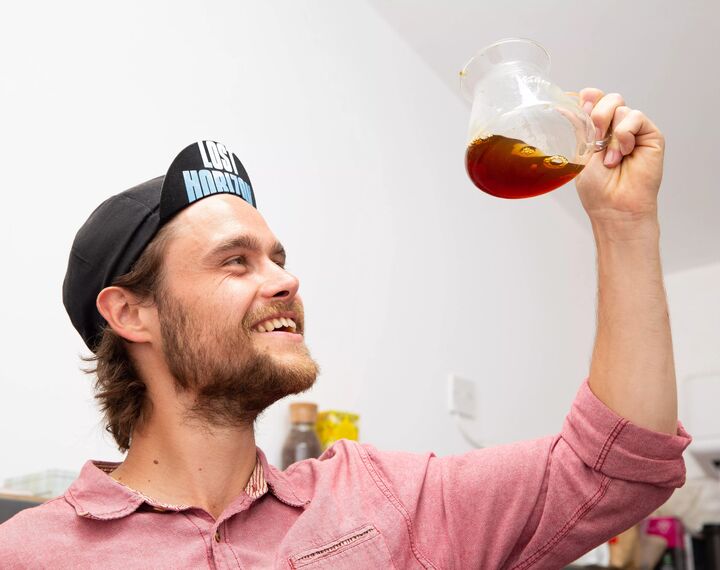 A man in a hat with the words 'Lost Horizon' on the lid holds up a glass with coffee in. He is looking up at the coffee and smiling.