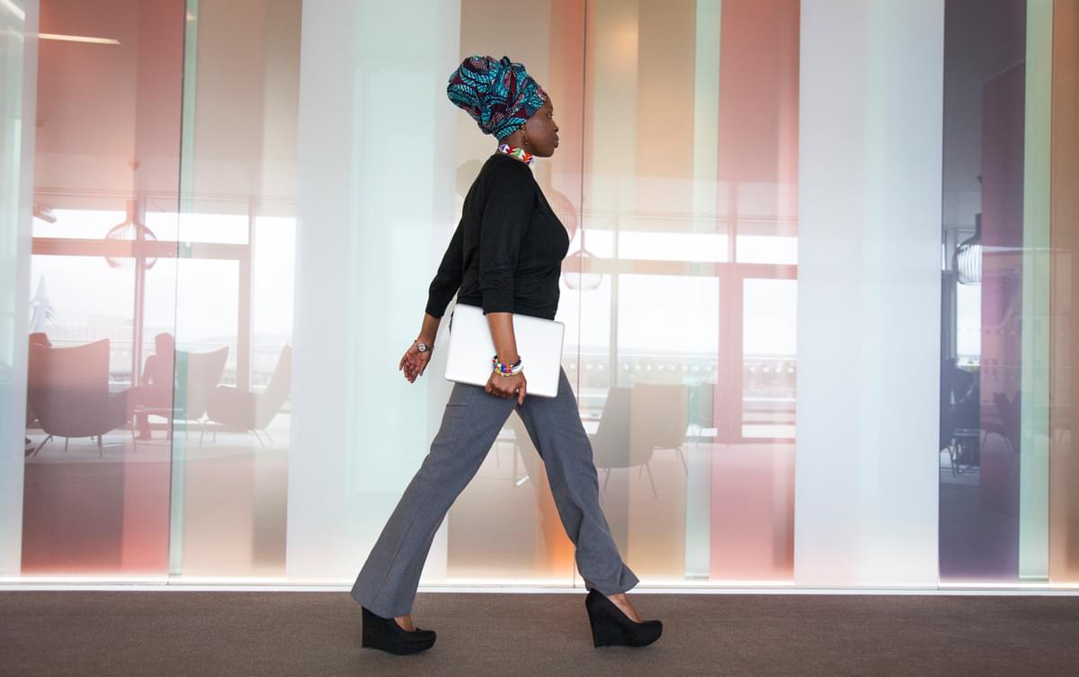 A black woman wearing a head wrap and carrying a laptop strides along a corridor. You can see office chairs reflected in the  colourful glossy wall behind her.