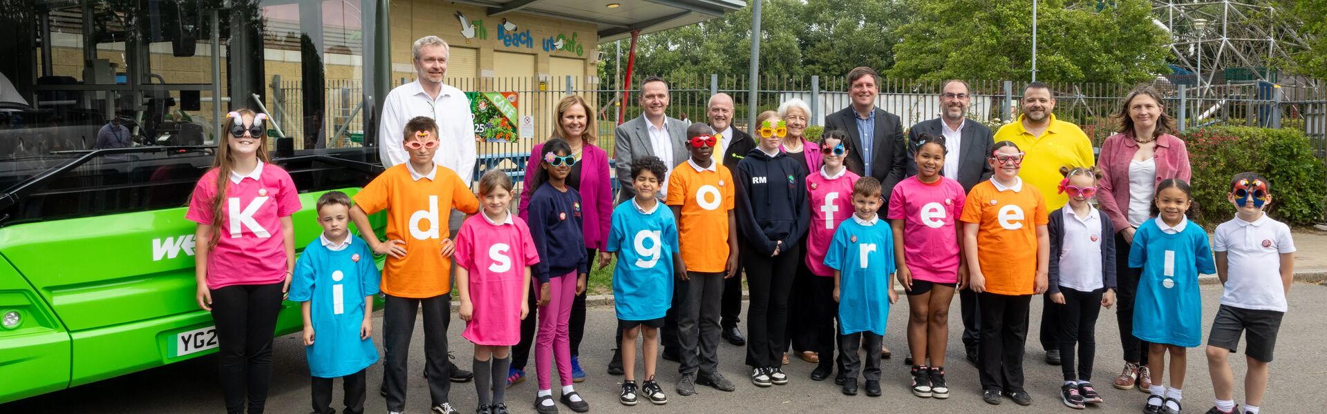 A group portrait of children in colourful t-shirts that spell out 'Kids Go Free!' accompanied by council leaders and Mayor Helen Godwin. The group is standing next to a green bus and in front of a play park.