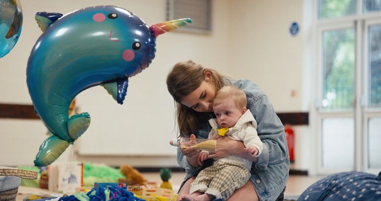 a mother holding her young baby and showing him a rattle. There is a large foil balloon next to them, in the shape of narwhal