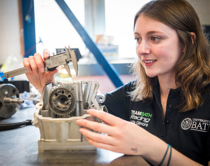 A female student is working on a silver item with cogs on, she is holding a metal spanner. She is wearing a black 'Team Bath Racing' polo top