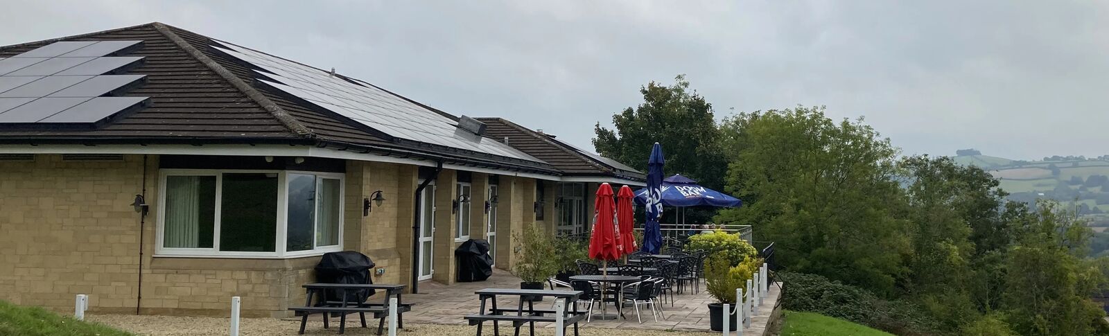 Golf course club house exterior with solar panels on roof and with umbrellas on patio and a view of hills behind 