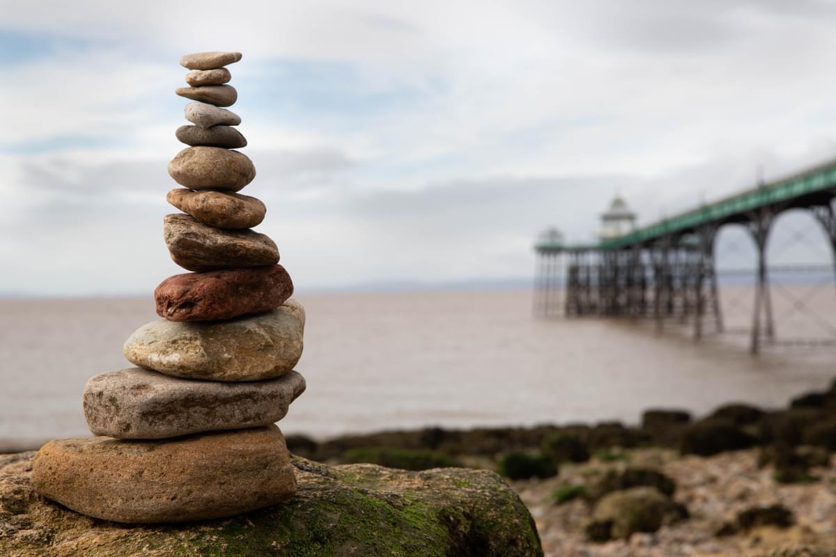 A stack of pebbles going from biggest to smallest is in the foreground. In the background is the water and a long pier and the stones of the beach.