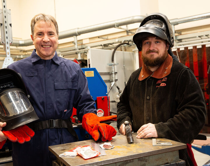 Metro Mayor Dan Norris is stood next to a young man at a welding course holding a hammer and the man is wearing a visor on his head. Dan is wearing an orange glove and they are both wearing boiler suits.