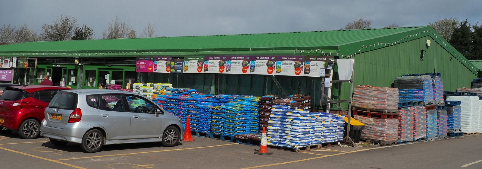 exterior view of a large green shed-like building, with bags of compost in front, and two parked cars