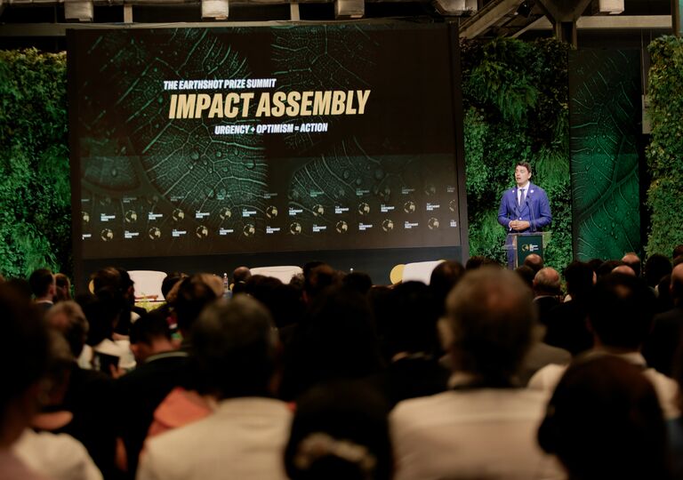 Man speaking in front of crowd, screen reads 