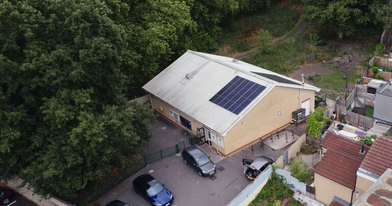 an aerial view of a large building with solar panels on the roof. there are cars parked outside, and lots of trees to one side of the building.