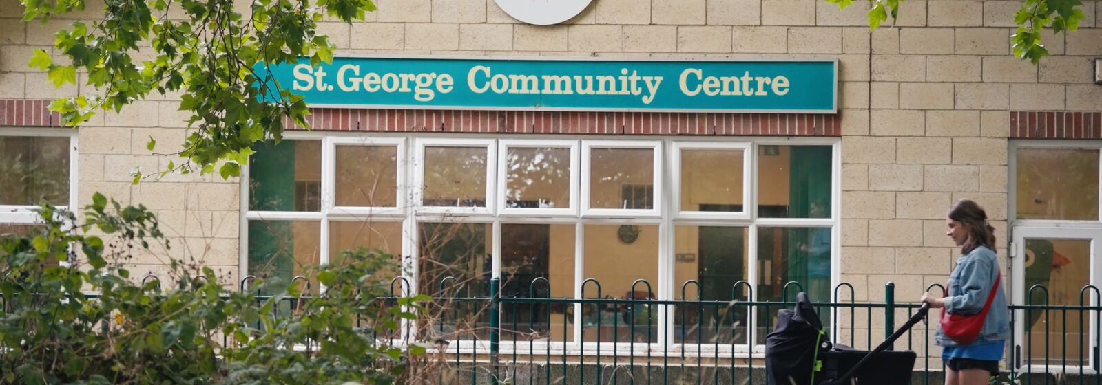 a young mother pushing a pushchair in front of a yellow brick building, with a teal sign that says 'St George Community Centre' in white writing.